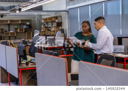 Diverse coworkers reviewing printed report at open-plan office desks with monitors, coffee cup Diverse coworkers reviewing printed report at open-plan office desks with monitors, coffee cup 140928344