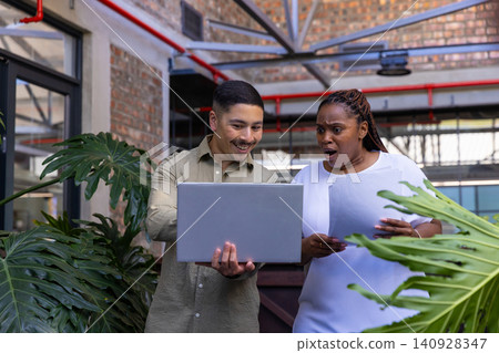 Diverse coworkers reviewing laptop and examining printed documents, tropical plants in office Diverse coworkers reviewing laptop and examining printed documents, tropical plants in office 140928347