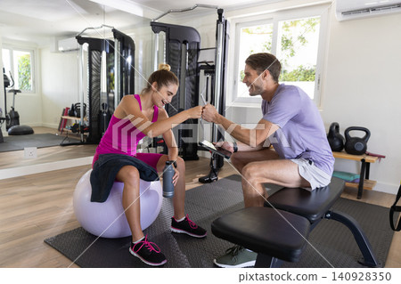 Trainer and client fist-bumping while sitting on stability ball holding water bottle in gym Trainer and client fist-bumping while sitting on stability ball holding water bottle in gym 140928350
