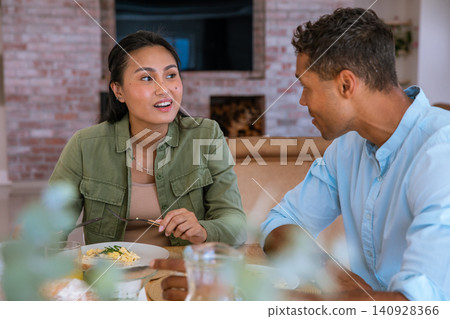 Diverse mid adult couple sharing pasta with fork at dining table in living room conversing Diverse mid adult couple sharing pasta with fork at dining table in living room conversing 140928366