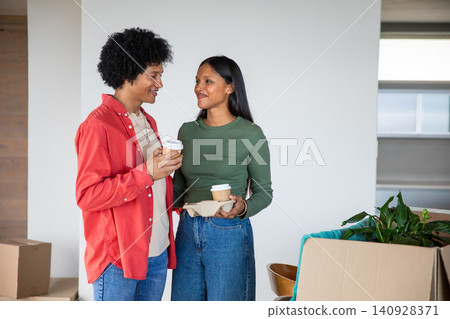 Diverse couple standing in room unpacking moving boxes, holding coffee tray near potted plant Diverse couple standing in room unpacking moving boxes, holding coffee tray near potted plant 140928371