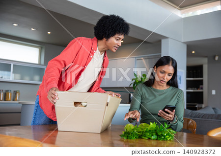 Diverse couple unpacking cardboard box and examining leafy greens, using smartphone in kitchen Diverse couple unpacking cardboard box and examining leafy greens, using smartphone in kitchen 140928372