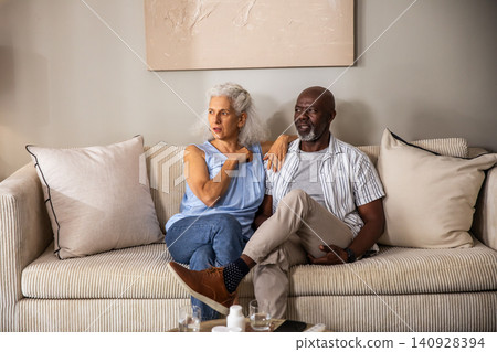 Diverse couple sitting on cream sofa in living room gesturing toward herself with painting behind Diverse couple sitting on cream sofa in living room gesturing toward herself with painting behind 140928394