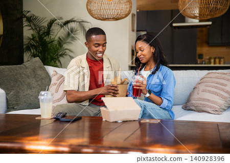 African American couple sharing takeout on sofa in living room with takeout boxes and clear cups African American couple sharing takeout on sofa in living room with takeout boxes and clear cups 140928396
