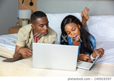 African American couple lying on bed at home pointing at open notebook while watching laptop screen African American couple lying on bed at home pointing at open notebook while watching laptop screen 140928397