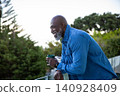 Senior african american man leaning on glass railing, green mug overlooking garden, copy space 140928409