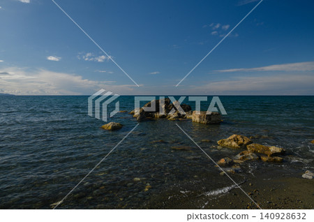 Serene coastline with gentle waves and rugged rocks under a clear blue sky at midday Serene coastline with gentle waves and rugged rocks under a clear blue sky at midday 140928632