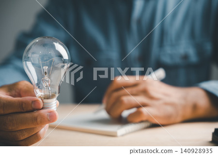 Hand holding a lightbulb, with a blurred background of a person writing in a notebook. Hand holding a lightbulb, with a blurred background of a person writing in a notebook. 140928935