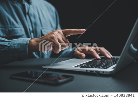 Close-up of a hand pointing at a laptop screen, with a smartphone in the foreground. The image is dark, highlighting the hand and the technology. Close-up of a hand pointing at a laptop screen, with a smartphone in the foreground. The image is dark, highlighting the hand and the technology. 140928941