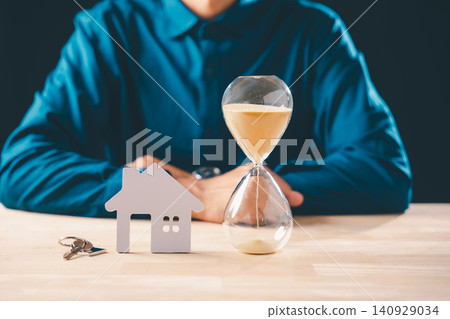 Person contemplates real estate property, hourglass shows time limit for home purchase, sand falling next to house model, keys for mortgage loan deadline Person contemplates real estate property, hourglass shows time limit for home purchase, sand falling next to house model, keys for mortgage loan deadline 140929034