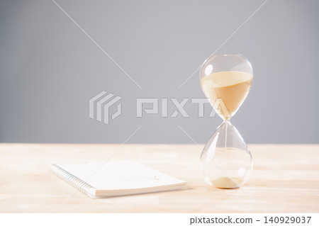 Hourglass time clock management concept displays sand flowing representing deadline countdown for business projects, spiral notebook waits on wooden table for notes Hourglass time clock management concept displays sand flowing representing deadline countdown for business projects, spiral notebook waits on wooden table for notes 140929037