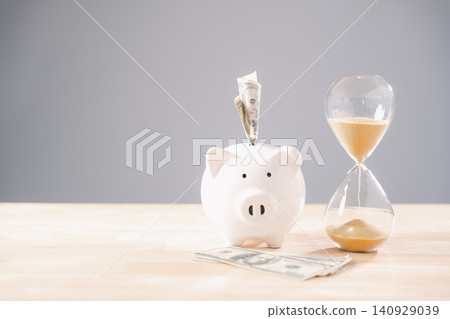 White piggy bank holds money, symbolizing saving money concept, time represented by elegant hourglass glass with sand flowing for business investment, future retirement planning White piggy bank holds money, symbolizing saving money concept, time represented by elegant hourglass glass with sand flowing for business investment, future retirement planning 140929039