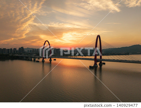 Aerial view captures golden sunset framing a prominent red arch bridge spanning a wide river with city skyline and mountains at dusk Aerial view captures golden sunset framing a prominent red arch bridge spanning a wide river with city skyline and mountains at dusk 140929477