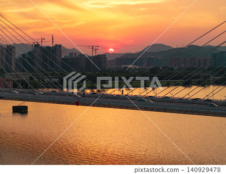 Aerial view captures a golden hour light bathes a modern city skyline and cable-stayed bridge spanning a calm river at dusk Aerial view captures a golden hour light bathes a modern city skyline and cable-stayed bridge spanning a calm river at dusk 140929478