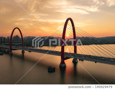 Aerial view captures golden sunset framing a prominent red arch bridge spanning a wide river with city skyline and mountains at dusk Aerial view captures golden sunset framing a prominent red arch bridge spanning a wide river with city skyline and mountains at dusk 140929479