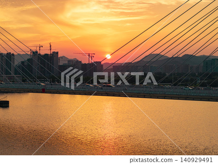 Aerial view captures a golden hour light bathes a modern city skyline and cable-stayed bridge spanning a calm river at dusk Aerial view captures a golden hour light bathes a modern city skyline and cable-stayed bridge spanning a calm river at dusk 140929491