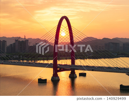 Aerial view captures golden sunset framing a prominent red arch bridge spanning a wide river with city skyline and mountains at dusk 140929499