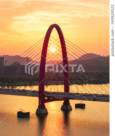 Aerial view captures vibrant red arch bridge spans a wide river as the sun sets, casting golden light on distant mountains and city skyline Aerial view captures vibrant red arch bridge spans a wide river as the sun sets, casting golden light on distant mountains and city skyline 140929522