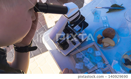 Curious boy looking through a microscope at an outdoor science fair. Enthusiastic kid exploring biology sample during an educational laboratory festival. Curious boy looking through a microscope at an outdoor science fair. Enthusiastic kid exploring biology sample during an educational laboratory festival. 140929690