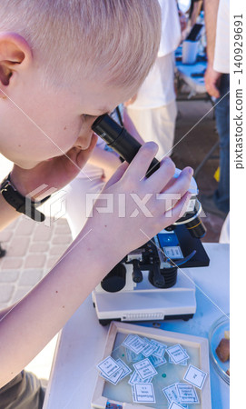 Curious boy looking through a microscope at an outdoor science fair. Enthusiastic kid exploring biology sample during an educational laboratory festival. 140929691