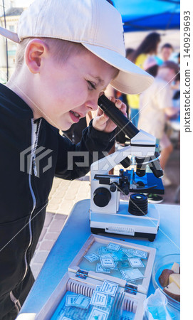 Curious boy looking through a microscope at an outdoor science fair. Enthusiastic kid exploring biology sample during an educational laboratory festival. Curious boy looking through a microscope at an outdoor science fair. Enthusiastic kid exploring biology sample during an educational laboratory festival. 140929693