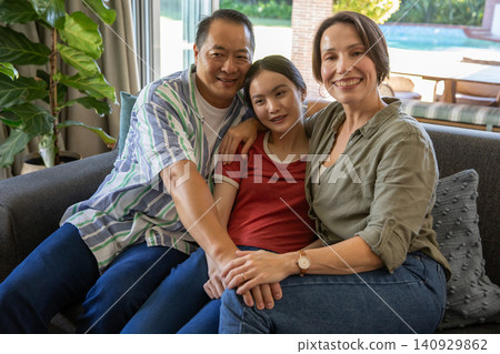 Asian family sitting closely together on gray sofa in living room, holding hands near sliding door 140929862