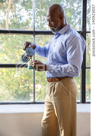 Senior African American man standing by metal window pouring coffee from French press in blue shirt Senior African American man standing by metal window pouring coffee from French press in blue shirt 140929887