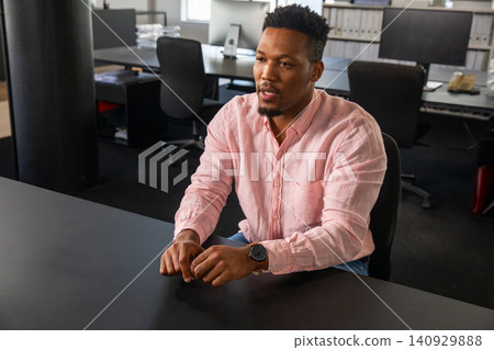 African American man speaking at large desk in open-plan office, wearing pink shirt and wristwatch 140929888