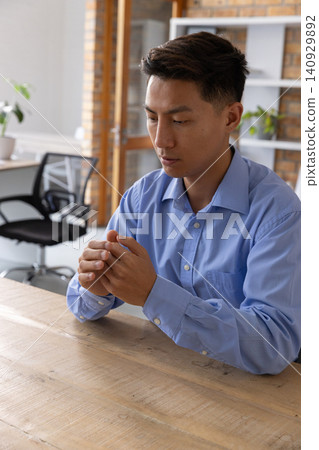 Asian man sitting at wooden table in office, wearing light blue shirt and clasping hands Asian man sitting at wooden table in office, wearing light blue shirt and clasping hands 140929892