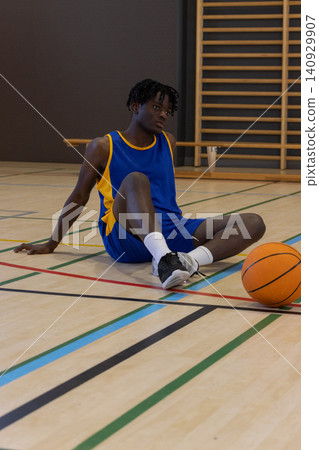 African American male basketball player sitting on court in blue kit holding ball near right foot African American male basketball player sitting on court in blue kit holding ball near right foot 140929907