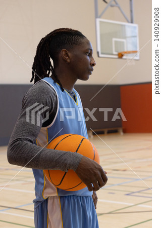 African American male holding basketball and facing right on gym hardwood court with hoop visible 140929908