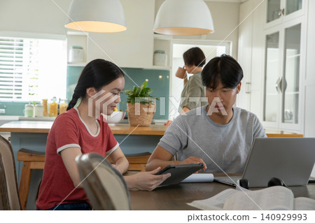 Asian family sitting at dining table in kitchen using tablet and laptop with notes, headphones, mug Asian family sitting at dining table in kitchen using tablet and laptop with notes, headphones, mug 140929993
