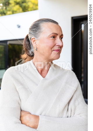Senior woman standing arms crossed at modern house entrance, wearing sweater and hoop earrings Senior woman standing arms crossed at modern house entrance, wearing sweater and hoop earrings 140929994