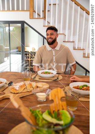 Bearded man in beige polo smiling while sitting at dining table with pasta, bread, salad bowl 140929997