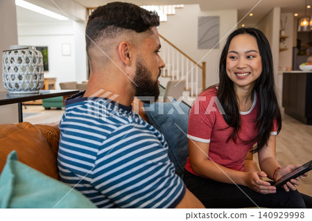 Couple sitting on leather sofa at home, Asian woman wearing red tee holding tablet, man listening Couple sitting on leather sofa at home, Asian woman wearing red tee holding tablet, man listening 140929998