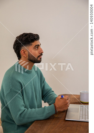 Indian man mid-20s wearing teal sweater sitting at table holding blue pen using laptop, copy space Indian man mid-20s wearing teal sweater sitting at table holding blue pen using laptop, copy space 140930004