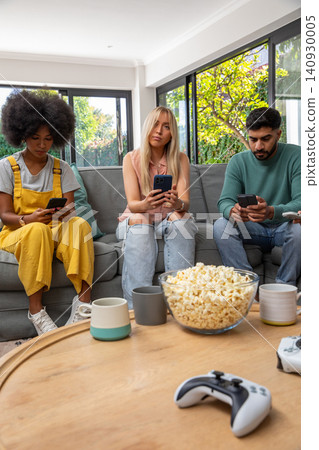 Diverse friends using smartphones on sofa in living room near popcorn bowl, one in yellow overalls Diverse friends using smartphones on sofa in living room near popcorn bowl, one in yellow overalls 140930005