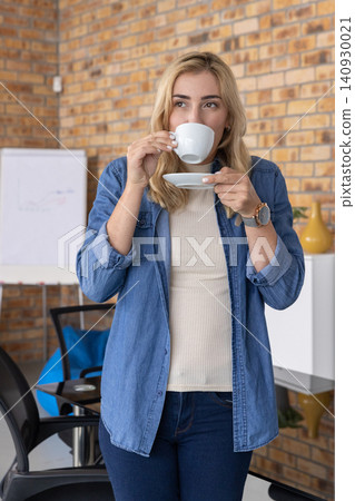 Mid-adult woman in denim shirt standing sipping from white cup holding saucer at office nook Mid-adult woman in denim shirt standing sipping from white cup holding saucer at office nook 140930021