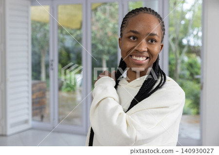 African American woman standing inside room by glass doors, wearing cream hoodie and showing braids African American woman standing inside room by glass doors, wearing cream hoodie and showing braids 140930027