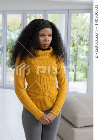 African American woman standing by sliding glass doors, wearing mustard sweater, gray sofa behind African American woman standing by sliding glass doors, wearing mustard sweater, gray sofa behind 140930033