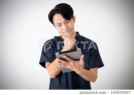 A middle-aged male doctor in navy blue scrubs looks troubled while looking at a tablet. A middle-aged male doctor in navy blue scrubs looks troubled while looking at a tablet. 140930047