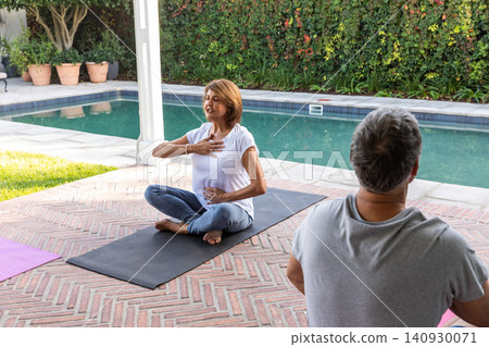 Senior couple practicing breathing on black yoga mat at backyard poolside patio with potted plants Senior couple practicing breathing on black yoga mat at backyard poolside patio with potted plants 140930071