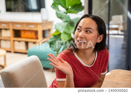 Asian adult female sitting at wooden table in cozy lounge wearing red T-shirt gesturing left hand Asian adult female sitting at wooden table in cozy lounge wearing red T-shirt gesturing left hand 140930082