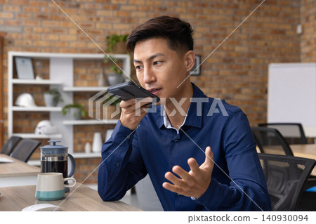 Asian man wearing navy shirt speaking into smartphone at desk with French press, copy space Asian man wearing navy shirt speaking into smartphone at desk with French press, copy space 140930094