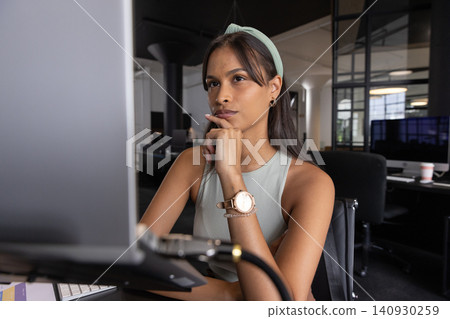 Adult asian woman sitting at desk in office, gazing at computer monitor with coffee cup 140930259