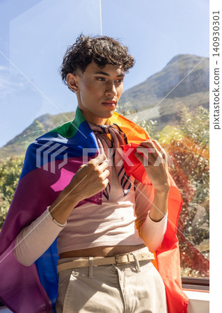 Asian man standing by window holding rainbow flag adjusting patterned scarf Asian man standing by window holding rainbow flag adjusting patterned scarf 140930301