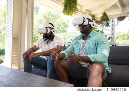 African american male friends using vr headsets and sitting on sofa under patio beams with plants African american male friends using vr headsets and sitting on sofa under patio beams with plants 140930326