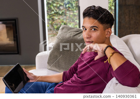 Man sitting on light grey sofa holding tablet in living room with fireplace insert, windows Man sitting on light grey sofa holding tablet in living room with fireplace insert, windows 140930361