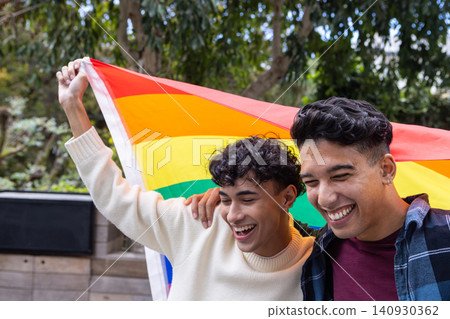 Two men standing on patio holding rainbow pride flag under leafy canopy and planter boxes 140930362