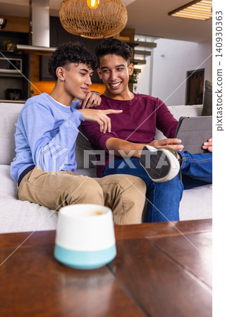 Two male friends sitting on sofa in living area pointing at tablet and smart speaker 140930363